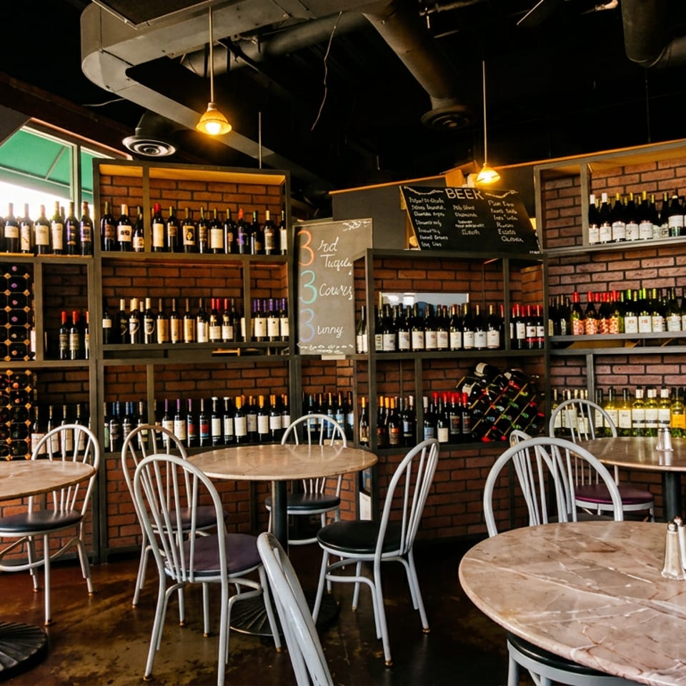 Rustic restaurant dining room with wooden tables, metal chairs, exposed brick walls, wine-filled shelves, and a beer chalkboard.