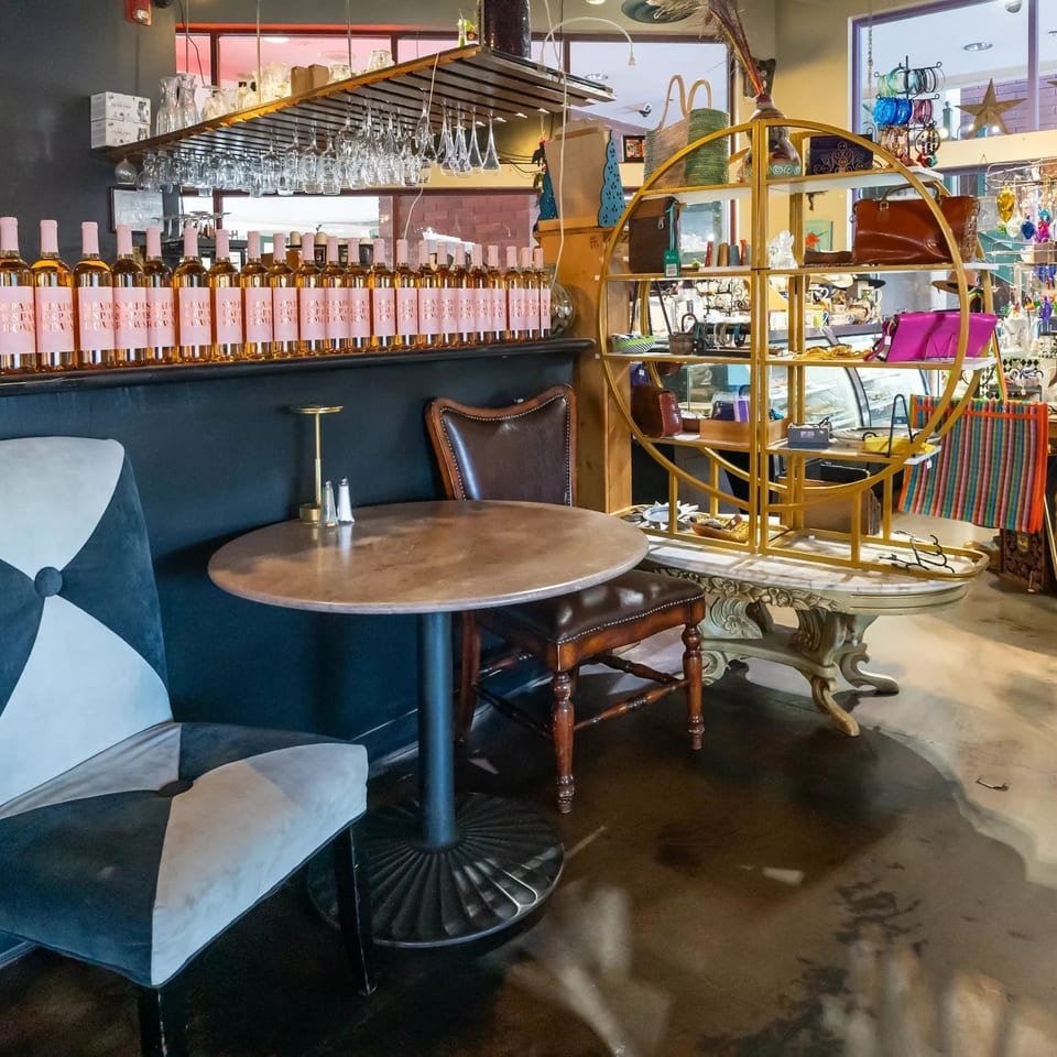 Eclectic restaurant interior with round tables, upholstered chairs, hanging glassware, wine bottles, and colorful retail displays.