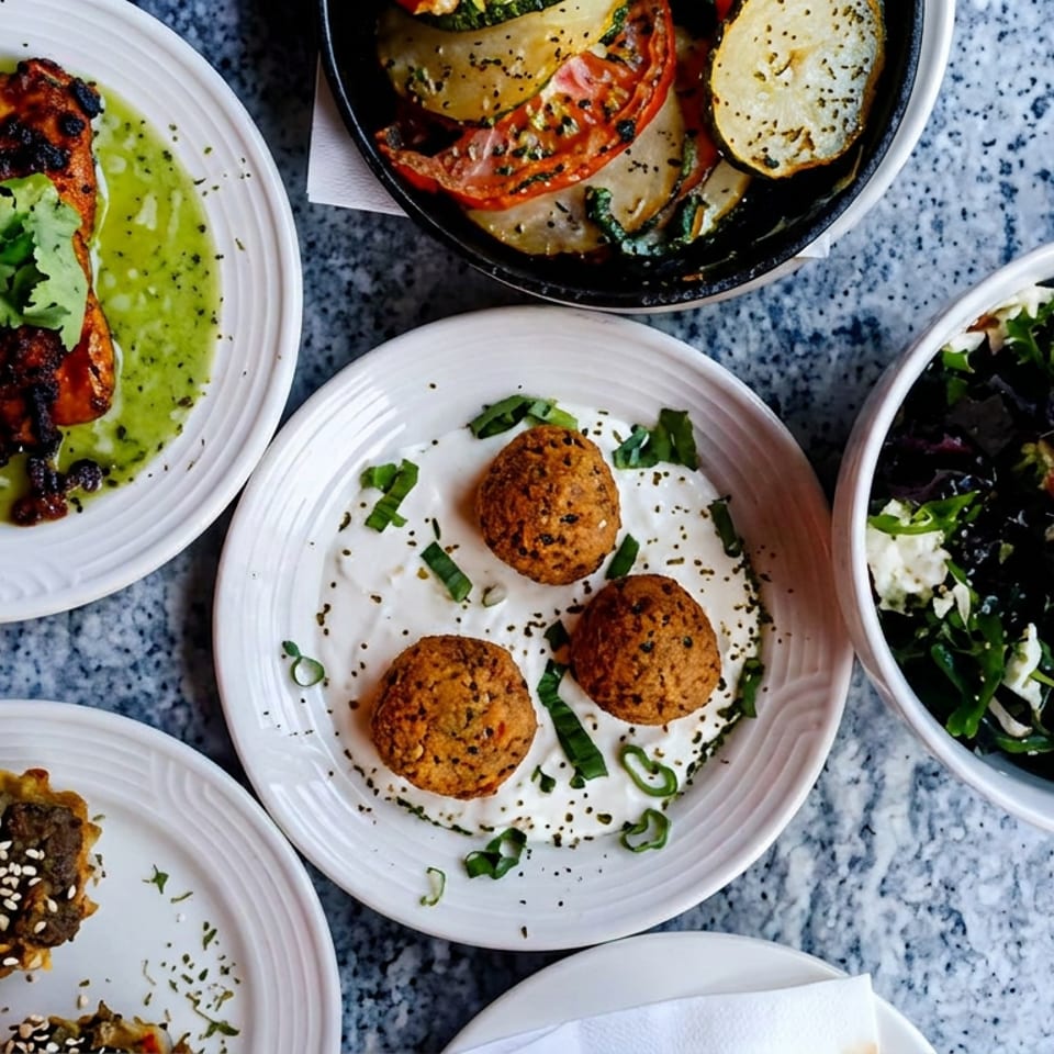 Overhead spread of colorful plated dishes, including grilled food with green sauce, roasted vegetables, falafel, salad, and sesame-topped bites.