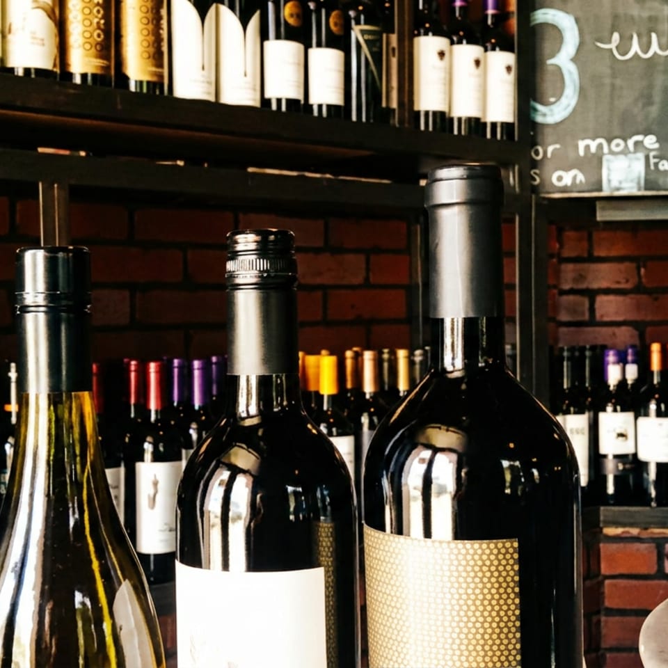 Three wine bottles in the foreground, surrounded by shelves of wine bottles against a brick wall and chalkboard.