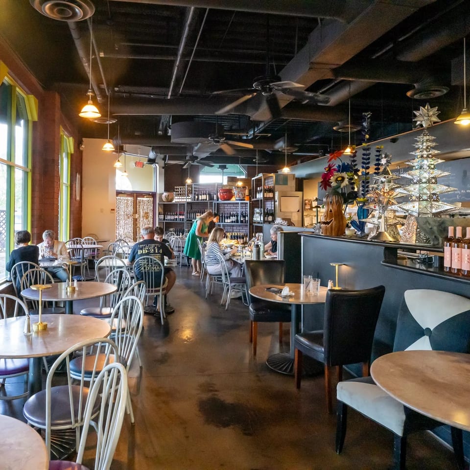 Bright restaurant dining room with round tables, seated diners, wine shelves, pendant lights, and a decorated bar area.