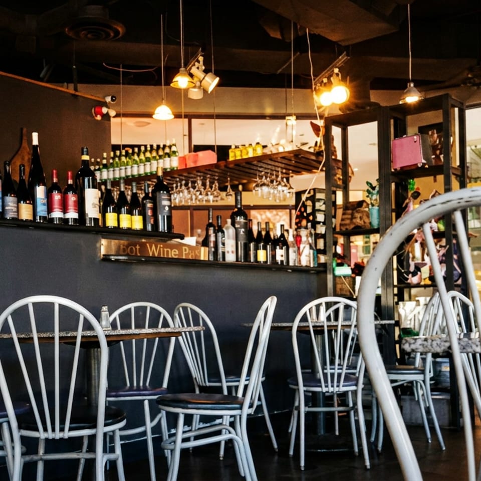 Warmly lit restaurant interior with white metal chairs, small tables, wine bottles, glassware, and eclectic decorative displays.